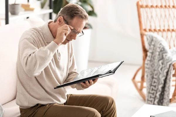 Senior Man Touching Glasses Holding Photo Album While Sitting Sofa — Stock Photo, Image