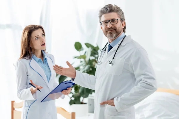Smiling Doctor White Coat Pointing Hand Shocked Colleague Looking Him — Stock Photo, Image
