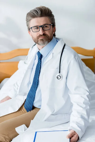 Όμορφος Γιατρός Λευκό Παλτό Κάθεται Στο Κρεβάτι Και Κοιτάζει Αλλού — Φωτογραφία Αρχείου