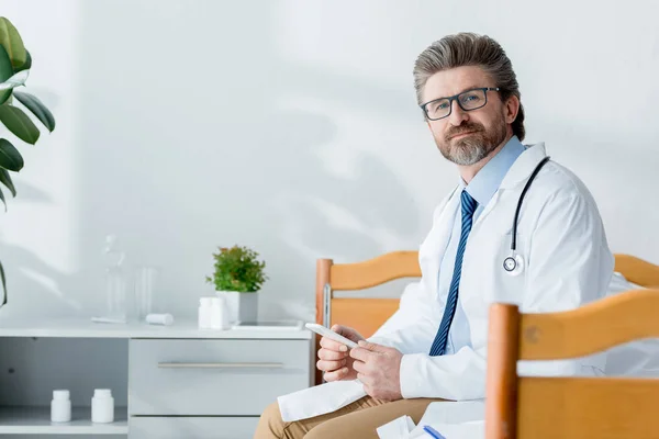 Handsome Doctor White Coat Sitting Bed Holding Smartphone Hospital — Stock Photo, Image