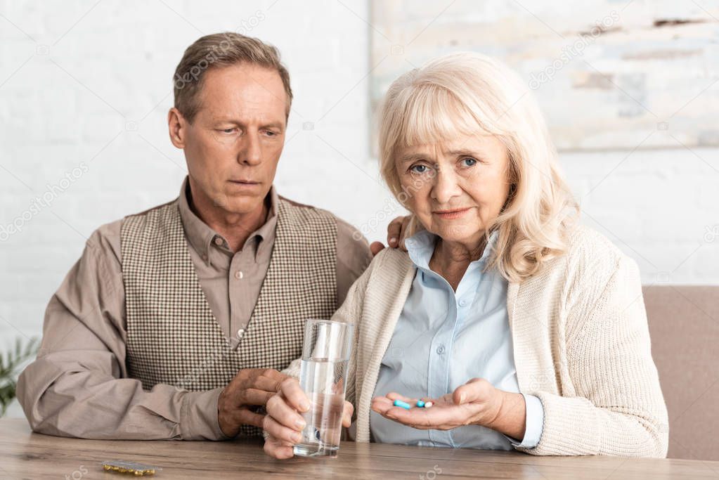 upset retired man sitting with sick wife holding glass of water and pills 