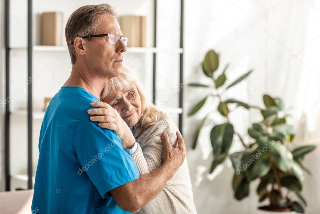 Upset senior woman with alzheimer hugging with doctor in glasses