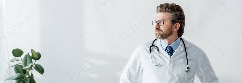 panoramic shot of handsome doctor in white coat looking away in hospital 