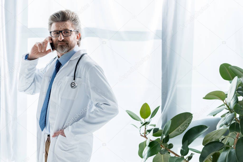 handsome doctor in white coat with hand in pocket talking on smartphone in hospital 