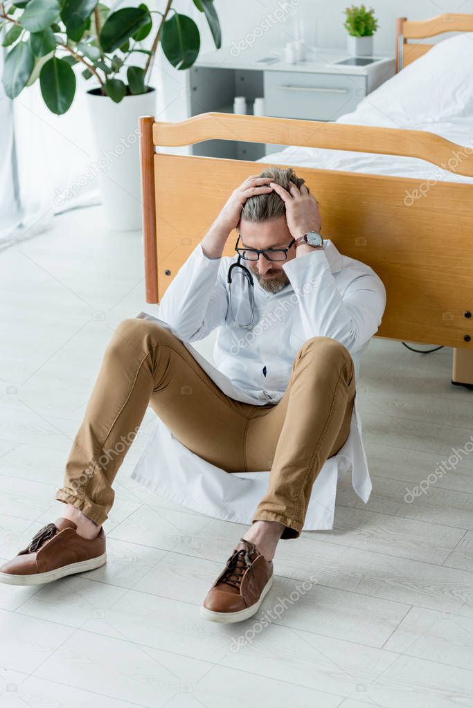 sad doctor in white coat sitting on floor and touching head in hospital 