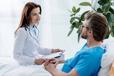Beyaz önlüklü doktor pano tutuyor ve hastanedeki hastaya bakıyor. 