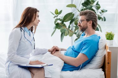 Beyaz önlüklü doktor el ele tutuşup hastanedeki hastaya bakıyor. 
