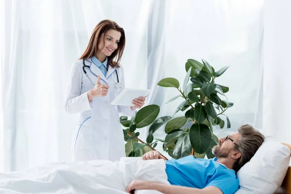 身穿白色外套 面带微笑的医生 手持数码平板电脑 躺在病床上 — 图库照片