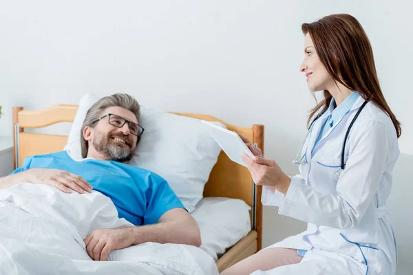 身穿白衣 面带微笑的医生 手持数码平板电脑 看着躺在病床上的病人 — 图库照片