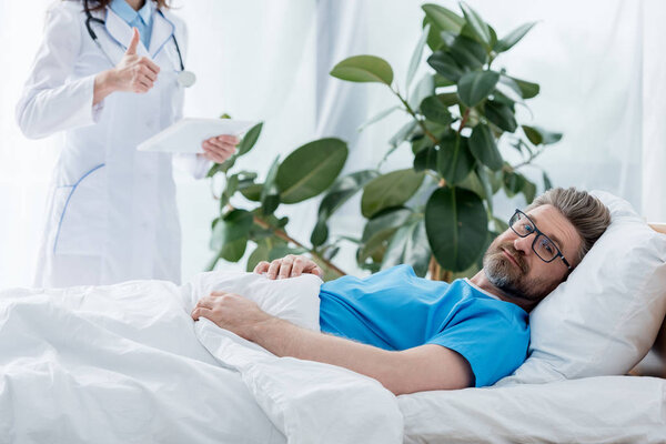 cropped view of doctor in white coat showing like and holding digital tablet and patient lying in bed in hospital 