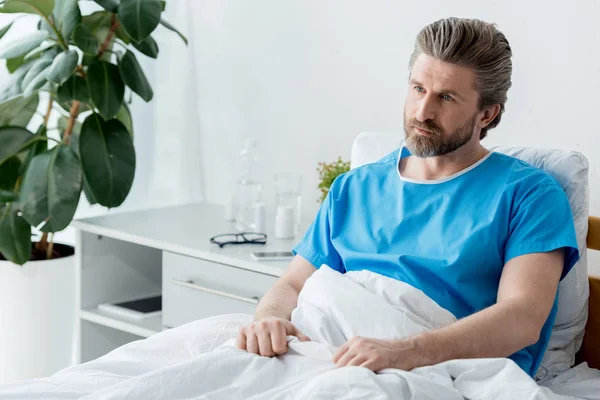 穿着医疗服的病人坐在床上 望着医院里的其他地方 — 图库照片
