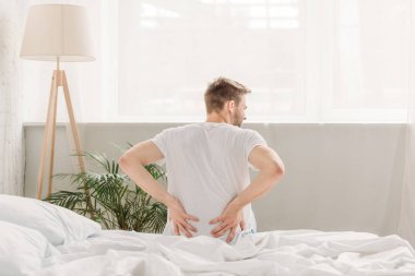 back view of young man sitting in bedroom and suffering from pain in loin clipart