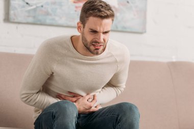 young man sitting on sofa and touching stomach while suffering from abdominal pain clipart