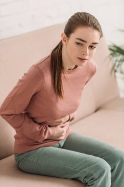 frowned girl sitting on sofa and suffering from stomach pain clipart