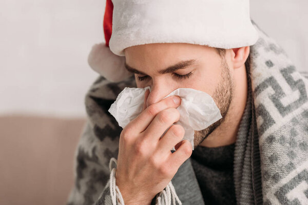diseased man in santa hat, wrapped in blanket, sneezing in napkin with closed eyes