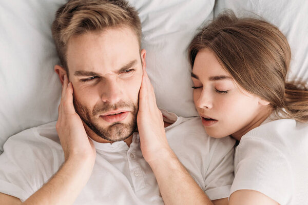 Top view of exhausted man plugging ears with hands while lying in bed near snoring wife
