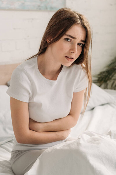 Нахмуренная девушка смотрит в камеру, страдая от боли в животе
