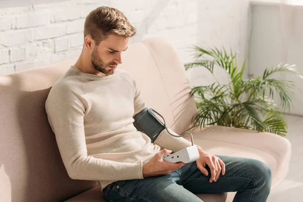 一个英俊的男人坐在家里的沙发上 用体能计测量血压 — 图库照片