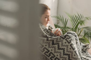 selective focus of ill girl covered with blanket and holding cup of warming drink clipart