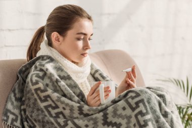 diseased girl, wrapped in blanket, looking at thermometer and holding cup of warming drink clipart