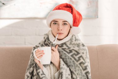 sick girl in santa hat, wrapped in blanket, looking at camera and holding cup of tea clipart