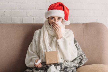 sick woman in santa hat sneezing in napkin and looking at camera 