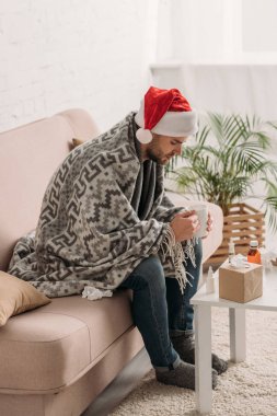 Noel Baba şapkalı hasta bir adam, battaniyeye sarılı, masanın yanında ilaçlarla oturuyor ve elinde bir fincan sıcak içecek tutuyor.