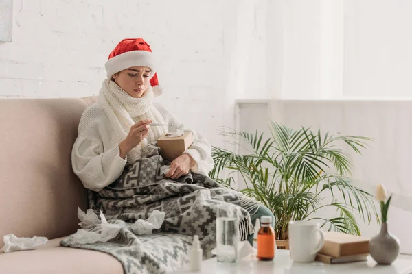 サンタ ハットの病気の女性は毛布に包まれ薬でテーブルの近くのソファに座り体温計を見て — ストック写真
