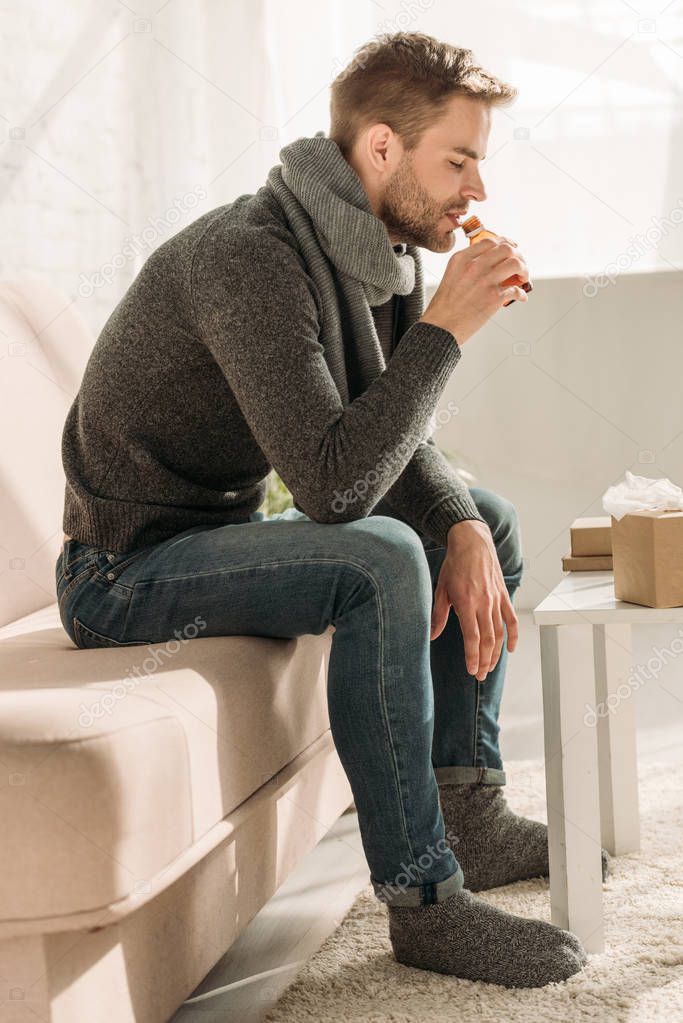sick man in warm scarf and socks sitting on sofa and drinking cough syrup