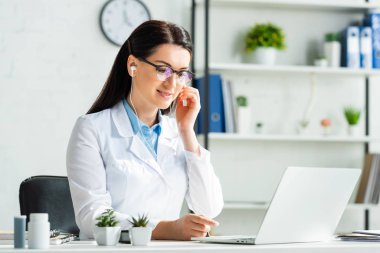 Kulaklıklı gülümseyen kadın doktor klinikteki laptopta hastayla çevrimiçi istişare yapıyor.