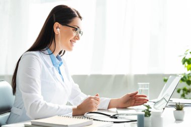 Kulaklıklı pozitif doktor klinikteki laptopta hastayla çevrimiçi istişare yapıyor.