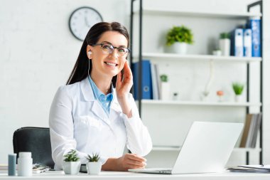 Kulaklıklı neşeli doktor, klinikteki laptopta hastayla çevrimiçi istişare yapıyor.