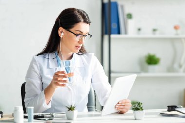 Ofisteki dijital tablette çevrimiçi danışmanlık yaparken elinde bir bardak su tutan kulaklıklı güzel doktor.
