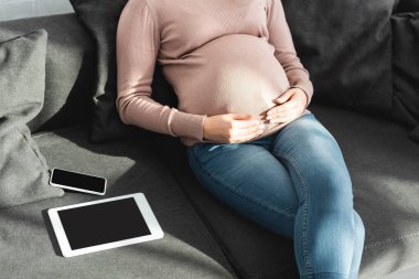 Dijital tablet ve akıllı telefonla koltukta otururken karnına dokunan hamile kadın görüntüsü.
