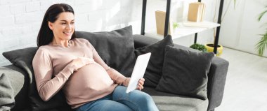 Evinde dijital tablet kullanan doktorla çevrimiçi istişare yapan hamile kadının panoramik görüntüsü.