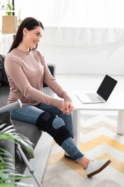 Kırık bacaklı gülümseyen bir kadın bilgisayarında doktorla internetten görüşme yapıyor.