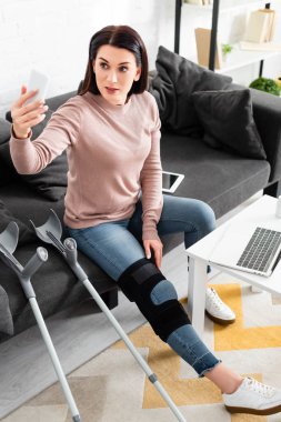 beautiful woman with broken leg having online consultation with doctor on laptop and smartphone