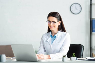 Bayan gülümseyen doktor, klinikte dizüstü bilgisayarla hastayla çevrimiçi görüşme yapıyor. 
