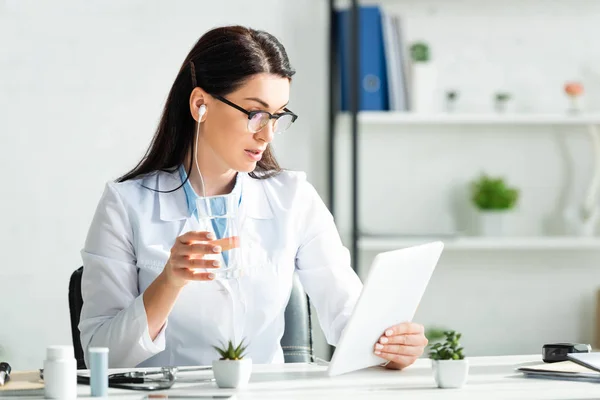 漂亮的医生 戴着耳机 一边拿着杯子喝水 一边在办公室里就数码平板电脑进行在线咨询 — 图库照片