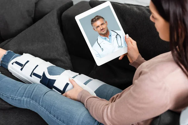 坐在沙发上与男医生在数码平板电脑上进行在线咨询的断腿妇女 — 图库照片