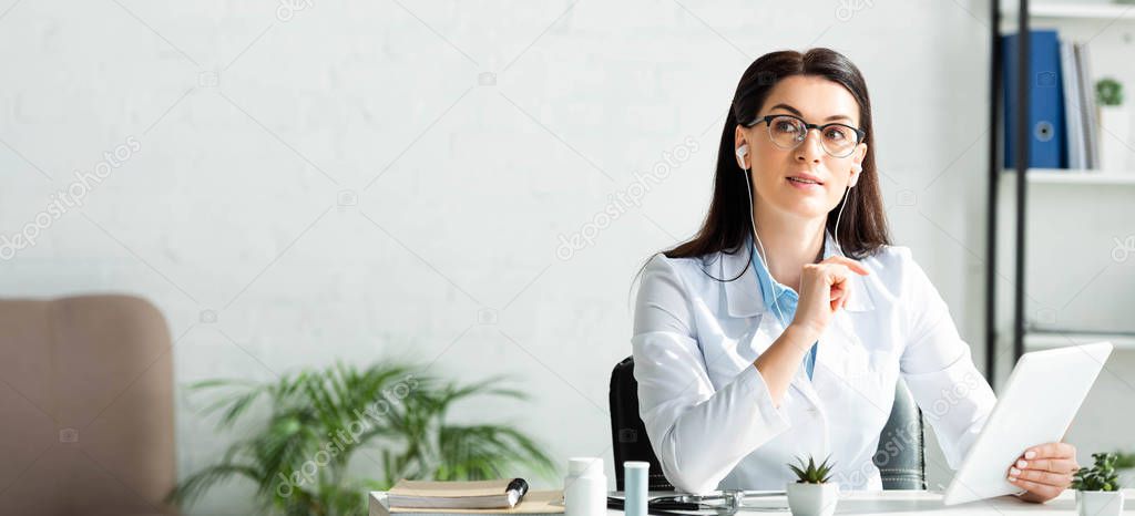 panoramic shot of thoughtful doctor in earphones having online consultation on digital tablet in office