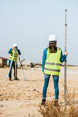 Selective focus of surveyors with survey ruler and digital level on construction site clipart