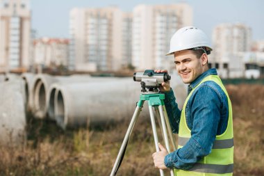 Surveyor with digital level smiling at camera with building materials at background clipart