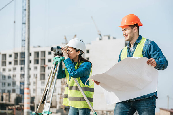 Smiling surveyor with blueprint and colleague with digital level
