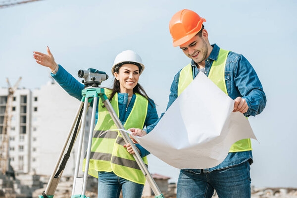 Smiling surveyor with digital level pointing away to worker with blueprint