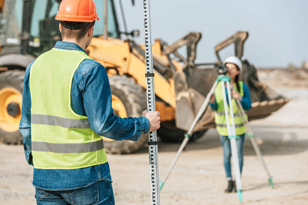 Selective focus of surveyors using ruler and digital level with bulldozer at background