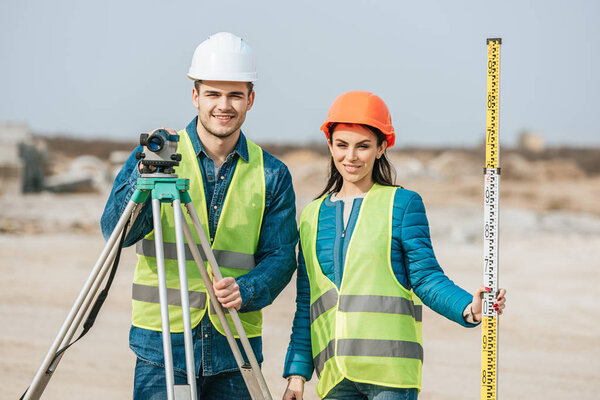 Smiling surveyors with ruler and digital level looking at camera