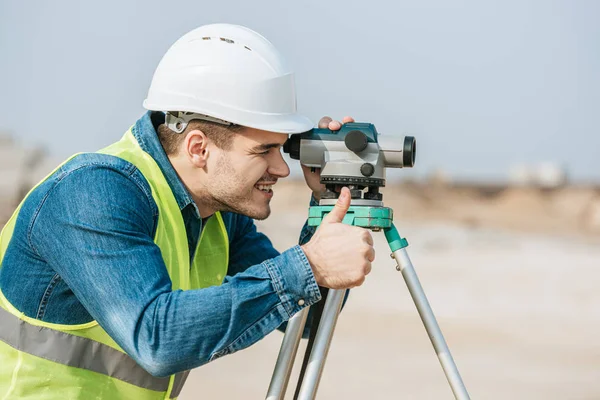 Sonriente Topógrafo Mirando Través Nivel Digital Mostrando Gesto Pulgar Hacia —  Fotos de Stock