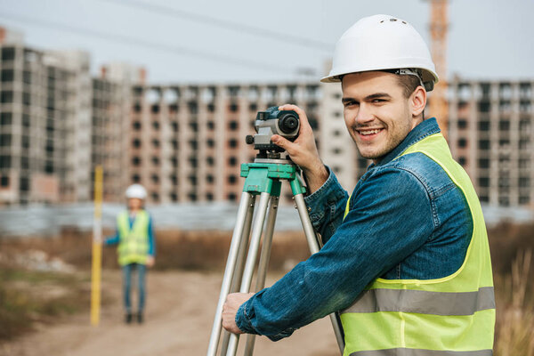 Selective focus of smiling surveyor with digital level and colleague at background