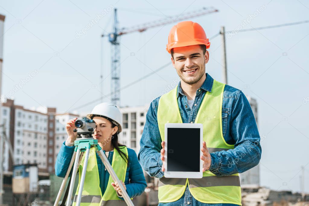 Smiling surveyor holding digital tablet with blank screen and colleague with measuring level at background